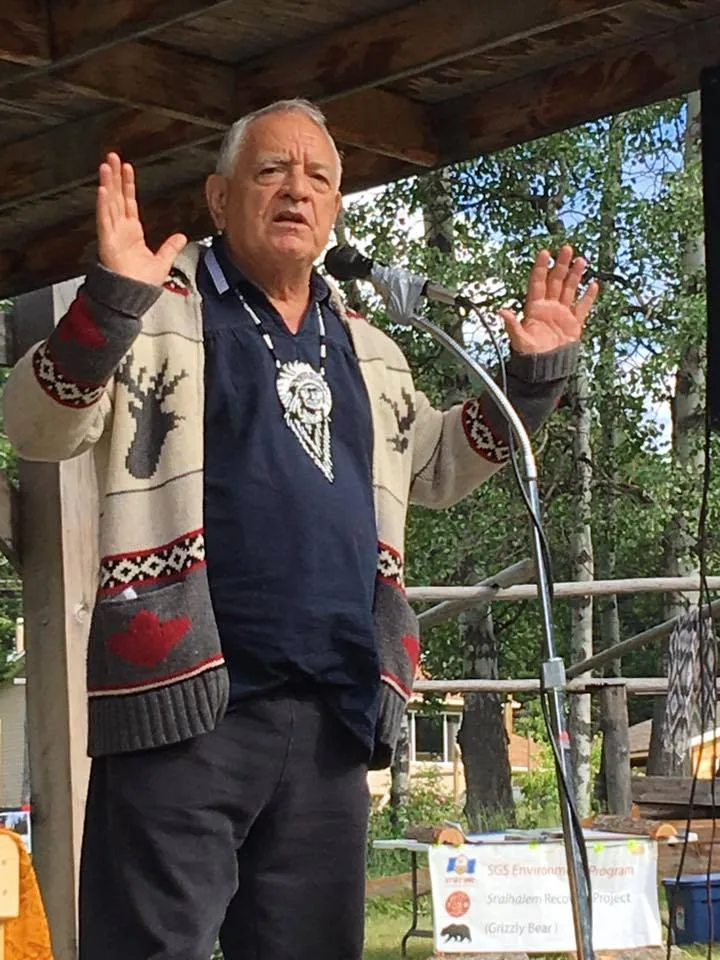 An elder with white hair, wearing a Cowichan-style sweater and a beaded medallion, speaks at an outdoor microphone with hands raised, beside a Grizzly Bear project sign.