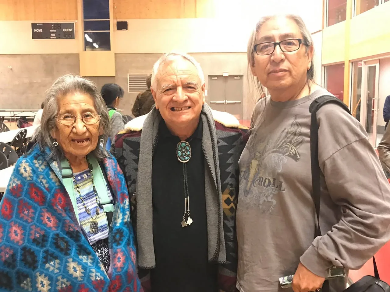 Three people stand together in a gymnasium: an elderly woman wrapped in a blue patterned blanket, a smiling white-haired man in a turquoise bolo tie, and a man with long gray hair and glasses.