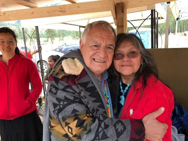 Phil Lane Jr., wrapped in a patterned blanket, embraces Mona Polacca, who wears a red jacket and turquoise necklace, under a wooden shelter as others stand nearby.