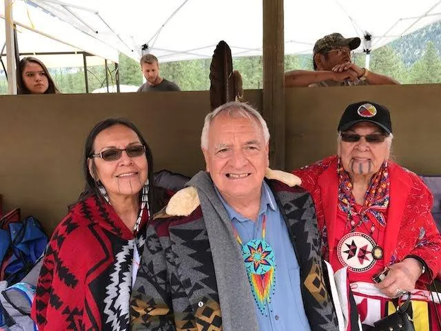 Phil Lane Jr., in a patterned blanket coat and beaded medallion, sits smiling between Unchi Agnes Baker Pilgrim, in red regalia and cap, and her daughter under a tent.