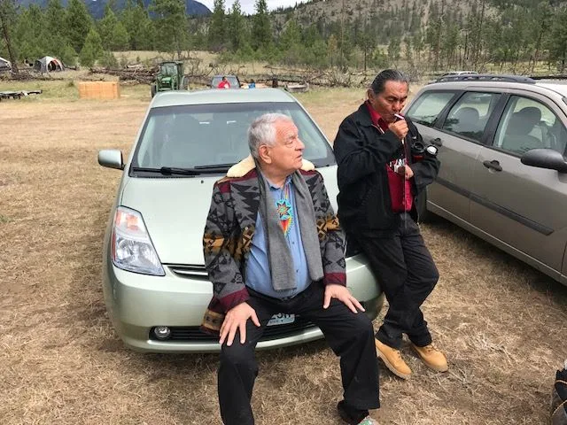 Phil Lane Jr. and Chief Darrell Bob rest against parked cars in a grassy field at Lillooet, with tents and forested hills behind them.