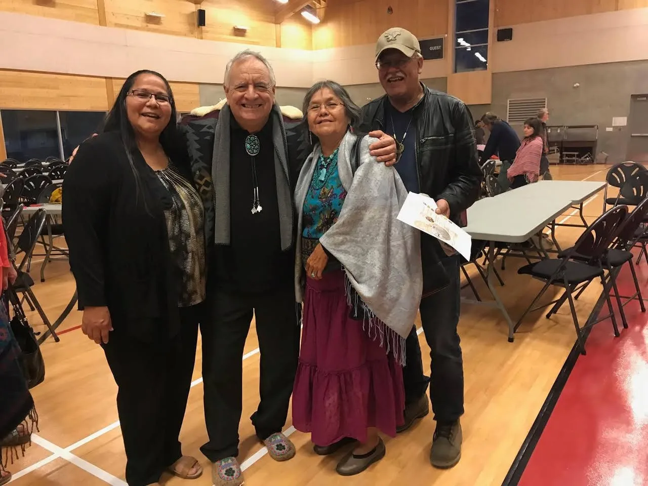 Four people smiling together in a gymnasium with tables and chairs: a woman in black, a white-haired man in a bolo tie and beaded moccasins, a woman in a silver shawl, and a man in a cap.