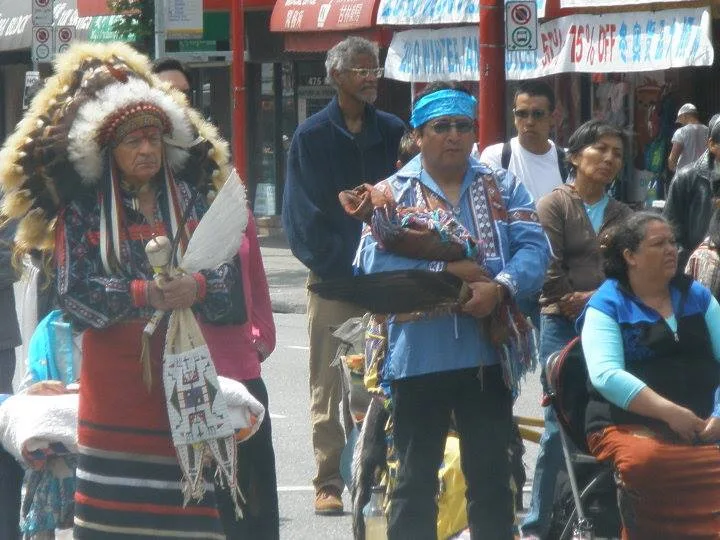 An elder in a full eagle-feather headdress holds a feather fan, beside a man in a blue headband cradling a bundle, among people gathered on a city street.