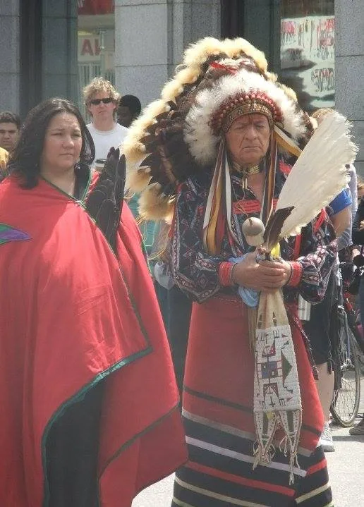 An elder in a feather headdress and beaded regalia holding a white feather fan stands beside a woman wrapped in a red blanket.