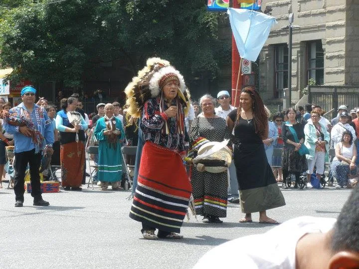 An elder in a feather headdress speaks into a microphone in the street, with women beside him and a crowd gathered behind.