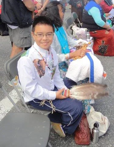 A smiling boy in glasses and a white buffalo shirt sits on a folding chair holding a feather fan, beside a seated woman.