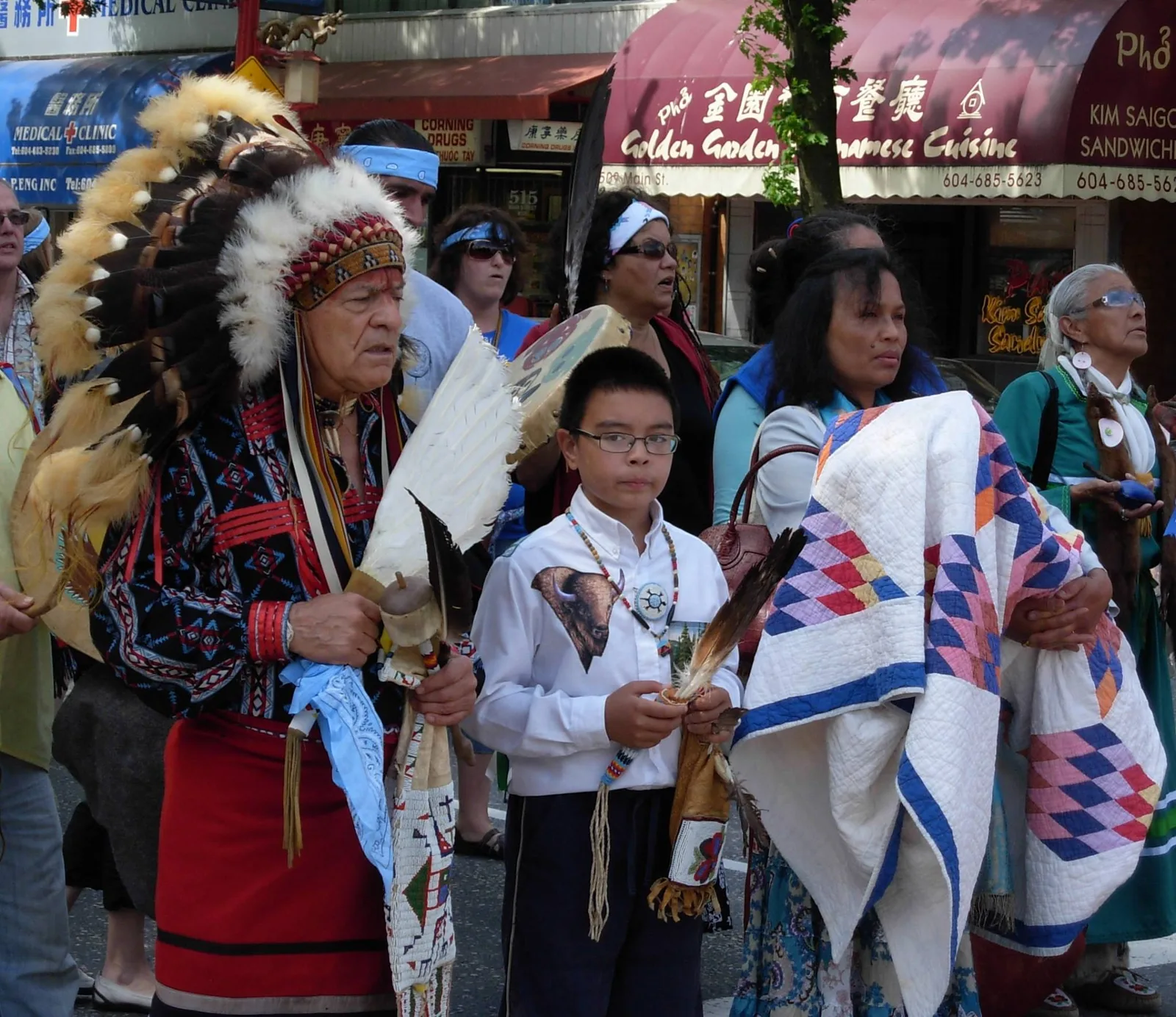 An elder in a feather headdress holding a feather fan walks with a boy in a white buffalo shirt and a woman carrying a star quilt.