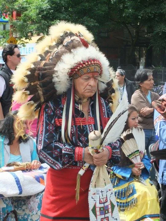 An elder in a feather headdress and patterned shirt stands with eyes lowered, holding a feather fan, as others gather around.