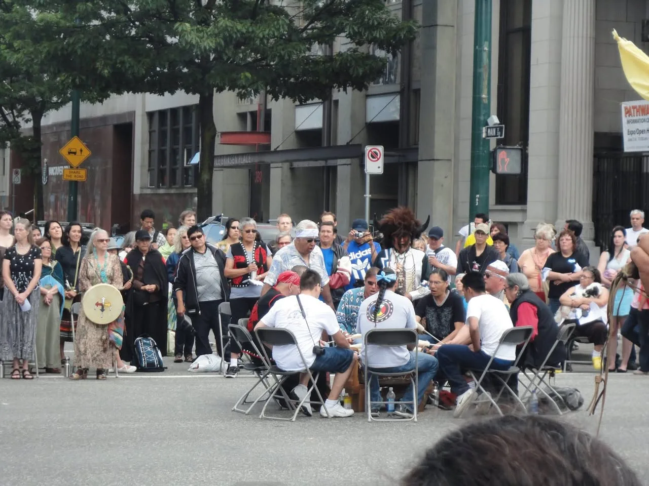A drum group seated in a circle sings in the middle of the street as a large crowd stands around them.