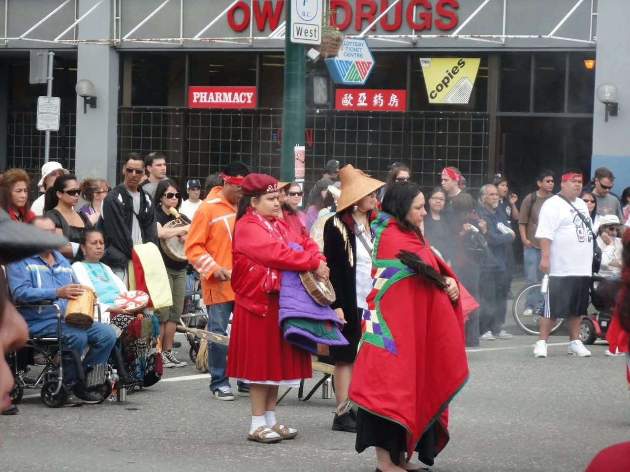 Women in red blankets and a woven cedar hat stand in the street holding drums, with a crowd and a pharmacy storefront behind.