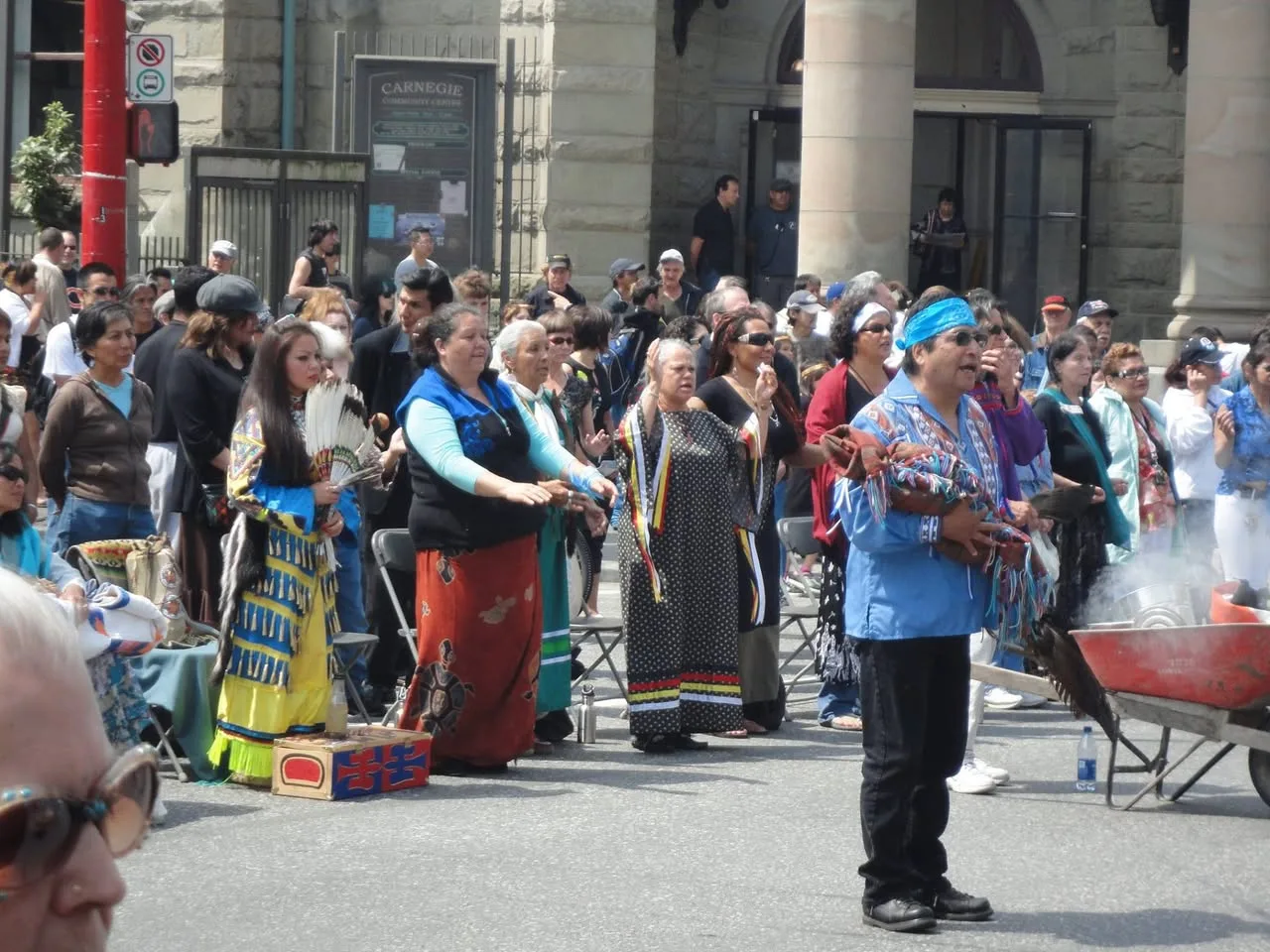 A man in a blue headband holds a bundle near a smudge smoke as women in regalia raise their hands in front of the Carnegie Community Centre.