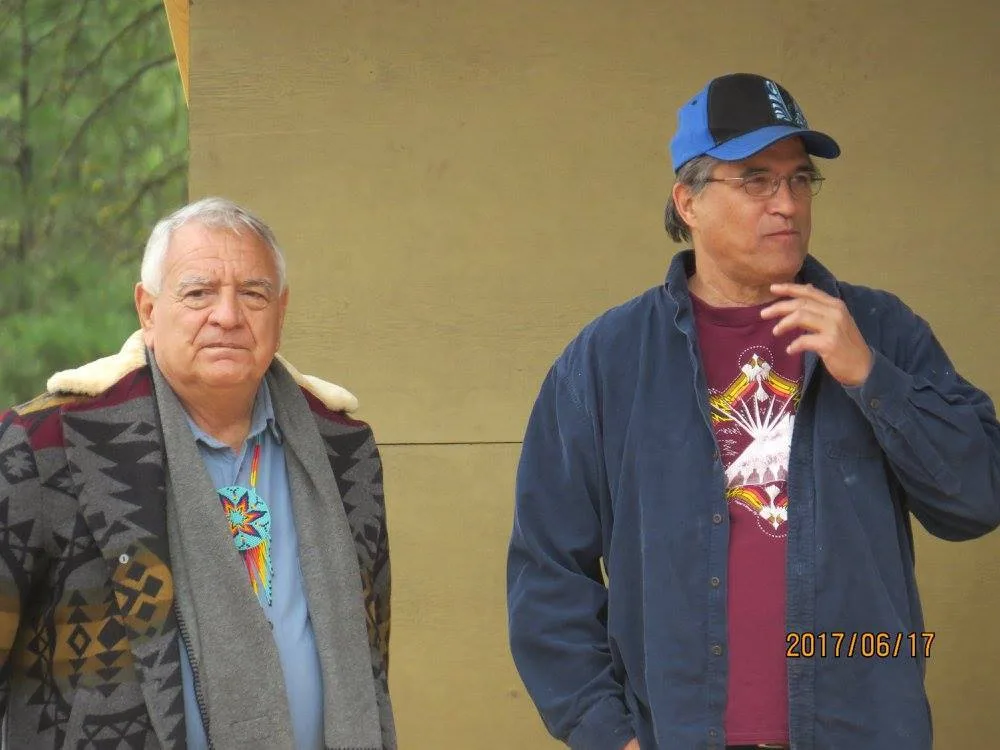 Two men standing outdoors: an older white-haired man in a patterned blanket coat with a beaded medallion, and a man in a blue cap and denim shirt over a maroon T-shirt.