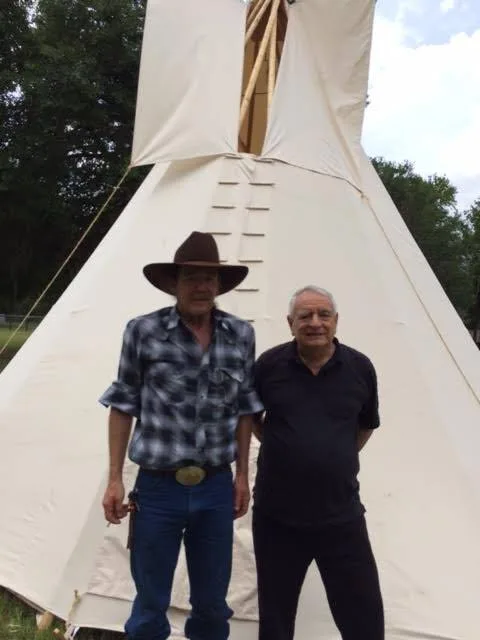 Two men stand in front of a tall white canvas tipi among trees, one in a brown cowboy hat, plaid shirt and jeans, the other gray-haired in a black shirt.