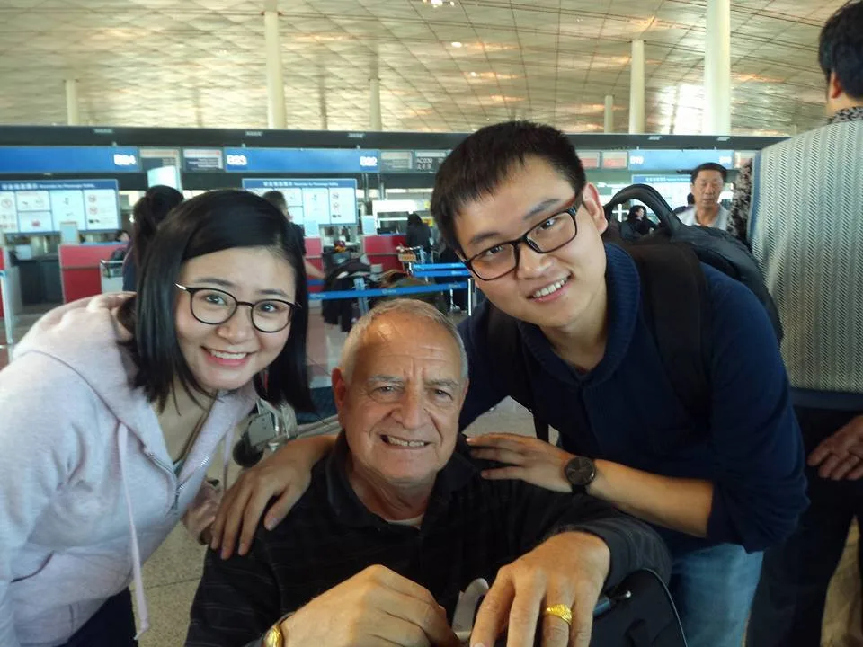 Phil Lane Jr., seated, smiles with a young woman and a young man in glasses, Sophia and Dan, in the check-in hall of Beijing Airport.