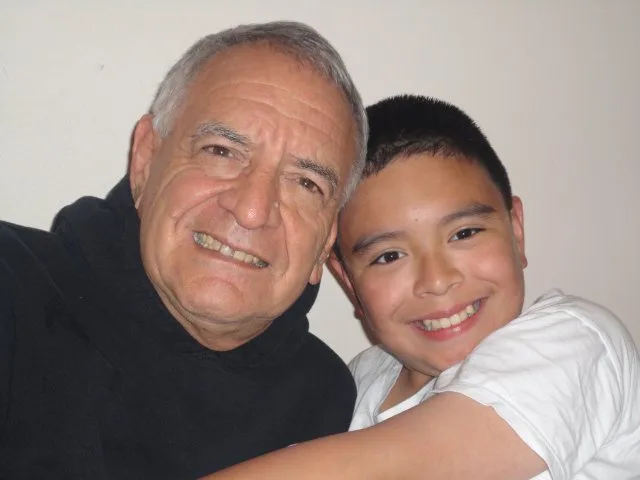 A smiling grey-haired man in a black hooded top sits cheek to cheek with a smiling boy in a white T-shirt who has an arm around him.