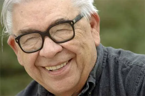 A smiling elderly man with white hair and black-framed glasses, wearing a dark denim shirt, photographed outdoors.