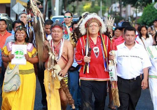 Indigenous delegates in traditional dress march down a Panama City street, led by an elder in a red shirt and feathered headdress holding a staff; Koda Darcy Demas, in dark glasses, walks behind.