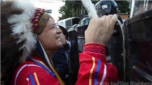 A smiling elder in a feather headdress and red ribbon shirt places his hand on a police riot shield as officers in helmets stand in a line.