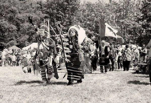Black-and-white photo of a powwow Grand Entry: dancers in feathered headdresses and regalia carry eagle staffs and flags across a grassy field as spectators watch.