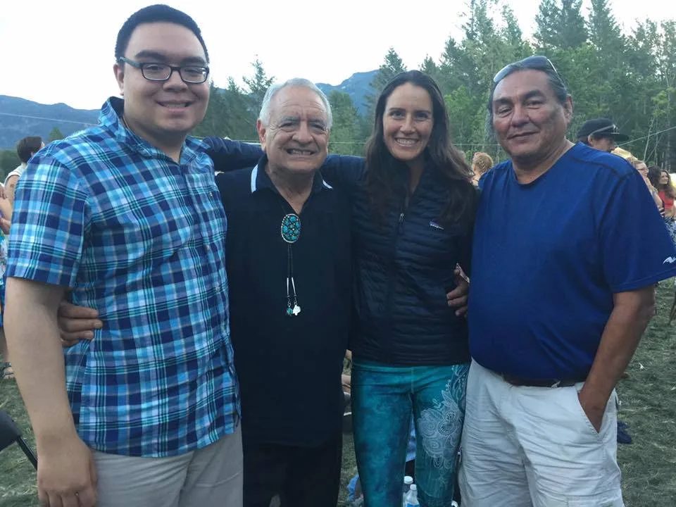 Four smiling people stand arm in arm outdoors on a grassy festival ground: a young man in glasses and a plaid shirt, an older man with a turquoise bolo tie, a woman, and a man in a blue shirt.