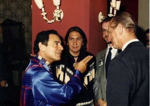 Phil Lane Jr., in a blue ribbon shirt, gestures as he speaks with Prince Philip, with Chief Leonard George in beaded regalia and Hugh Locke in glasses beside them.
