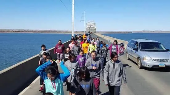 A large group of young people in hoodies walks across a long bridge over a wide river on a sunny day, with a minivan driving slowly alongside them.