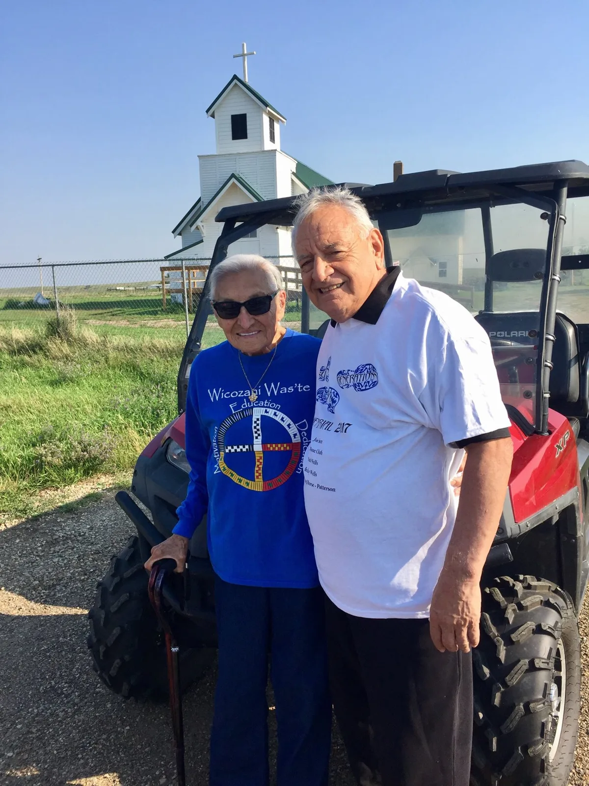 Auntie Hazel Red Bird, an elder in sunglasses and a blue Wicozani Was'te shirt holding a cane, stands with Phil Lane Jr. in front of a red utility vehicle and a white country church.