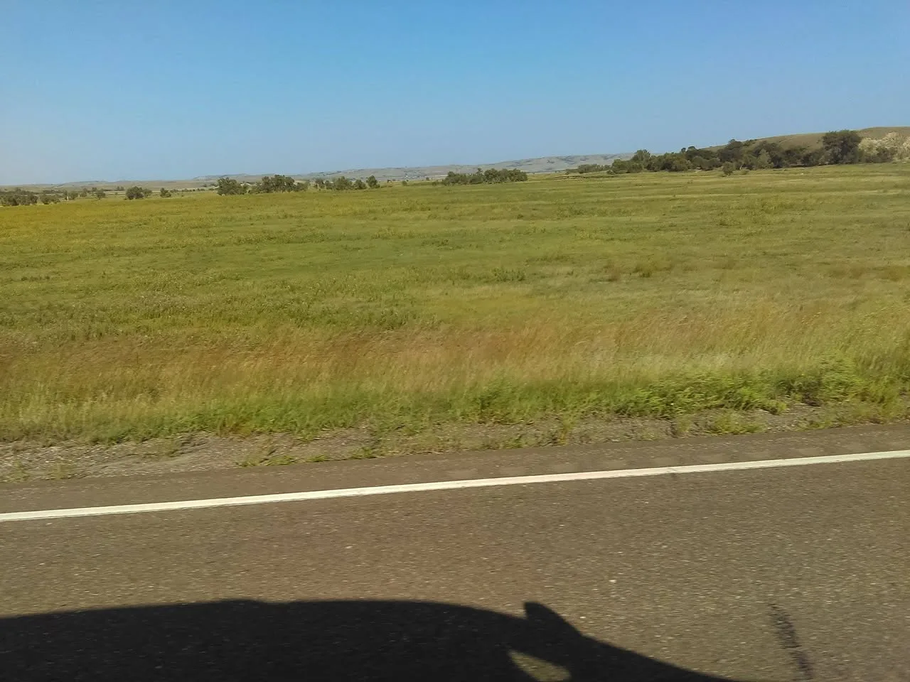 Wide grassy prairie stretching to distant hills and scattered trees, seen from a moving vehicle whose shadow falls on the road.