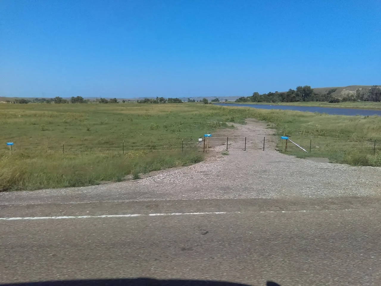 A gravel track leads from the highway through a wire fence gate toward open grassland beside a calm river under a clear blue sky.