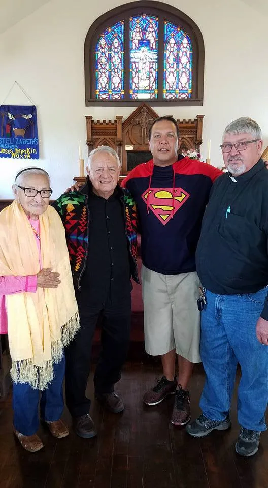 Four people stand together before the altar and stained glass window of St. Elizabeth's Church: an elderly woman in a yellow shawl, an older man in a patterned jacket, a man in a Superman hoodie, and a minister.