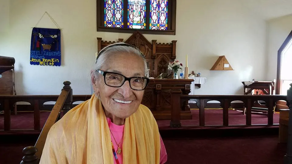 An elderly woman in glasses and a yellow shawl smiles warmly inside St. Elizabeth's Church, with the altar, stained glass and a blue banner reading "St. Elizabeth's wishes Jesus Tonpi Kin Waste" behind her.