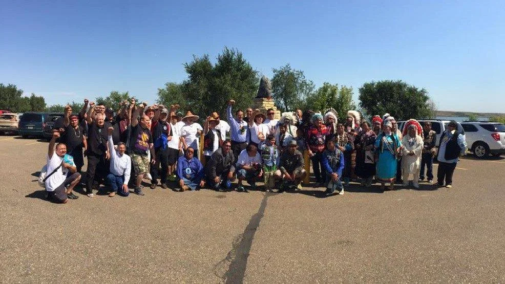 A large group of Indigenous leaders and supporters, some in feather headdresses and regalia, pose in a parking lot with raised fists beside a monument under a clear blue sky.