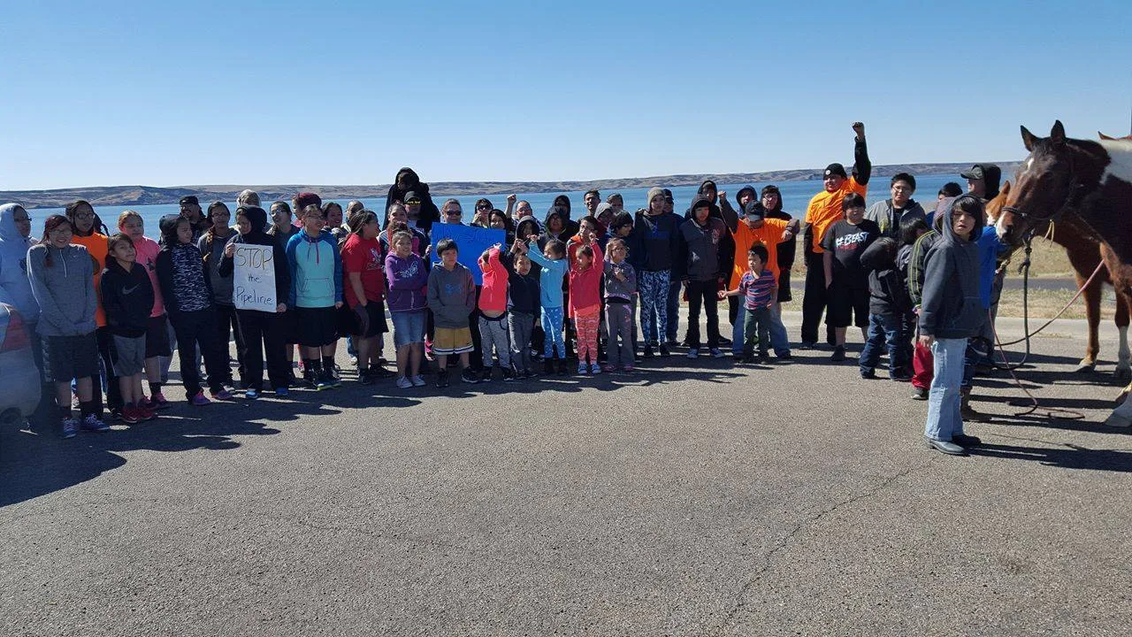 A large group of children, youth and adults stands on pavement before a wide lake under a clear blue sky; one child holds a sign reading "Stop the Pipeline" and a horse stands at right.