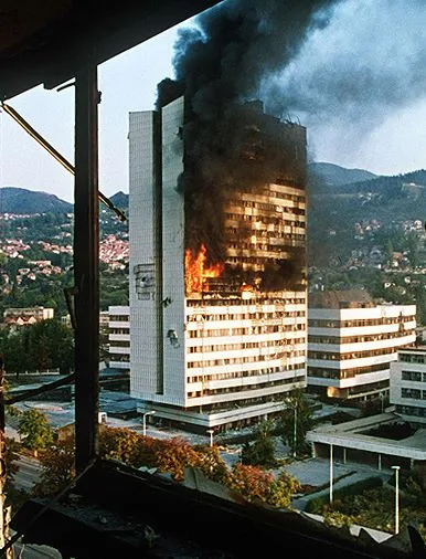 A tall office tower in Sarajevo burning, with flames on its lower floors and thick black smoke rising, seen through a damaged window frame with green hills behind.