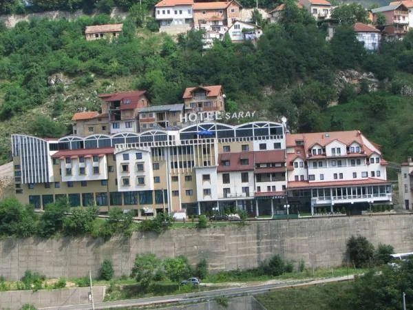 A large multi-story building with red-tiled roofs and a sign reading "Hotel Saraj" set on a green hillside above a retaining wall, with houses on the slope above.