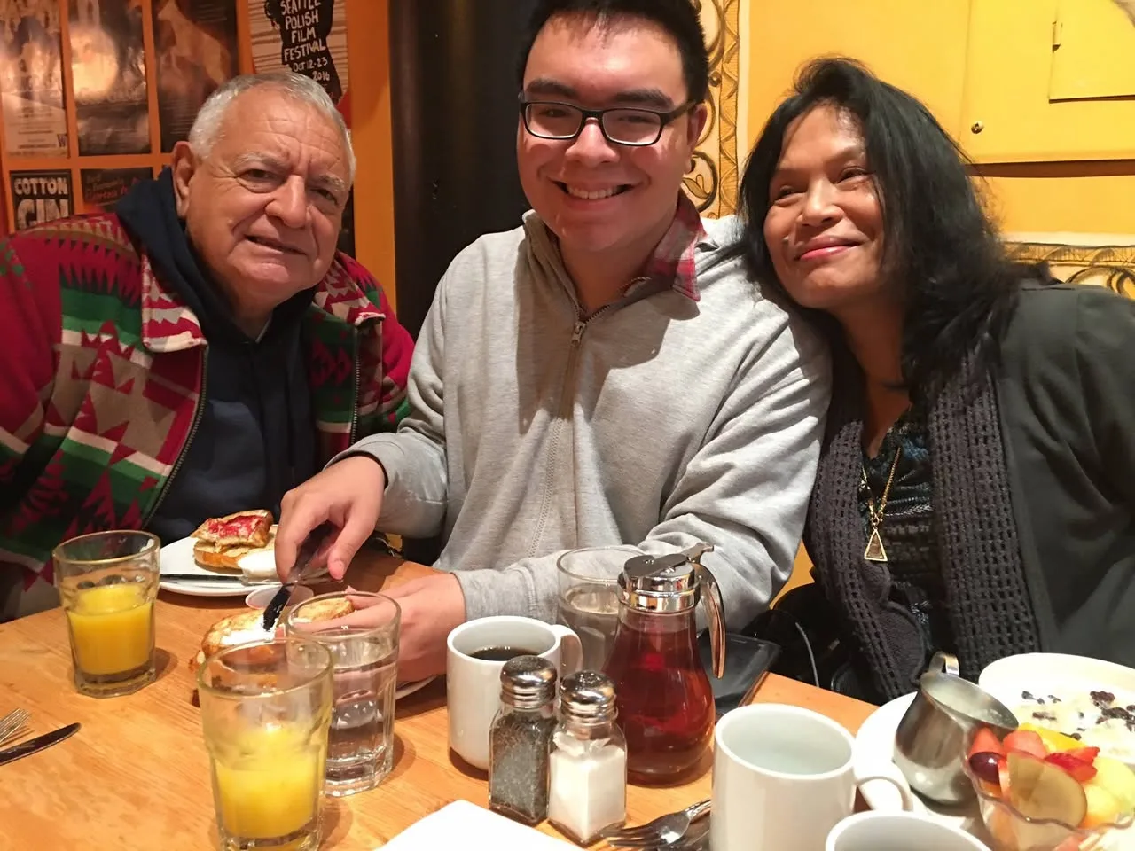 An older man in a red and green patterned jacket, a smiling young man in glasses and a grey sweater, and a woman with long dark hair share breakfast at a café table.