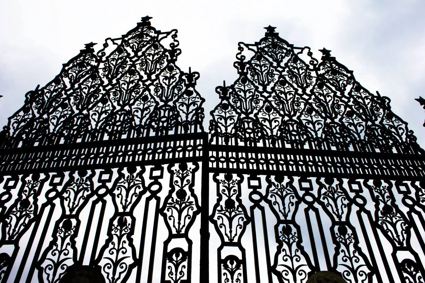 Ornate black wrought-iron gates with scrolling floral patterns and star-topped peaks, seen from below in silhouette against a pale cloudy sky.
