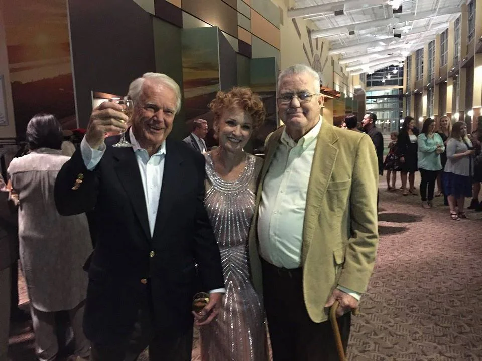 Three people at an evening reception: a white-haired man raising a glass, a woman in a sparkling silver gown, and a man in a tan jacket holding a cane. They are Greg's family, including brother Gary and sister Serita.