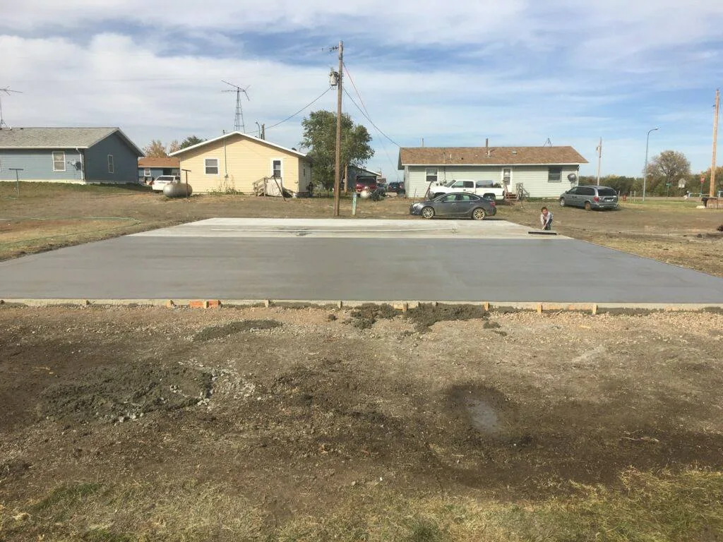 A freshly poured concrete slab for a basketball court on open ground, with a child near the far edge and houses, cars and power lines behind under a cloudy sky.