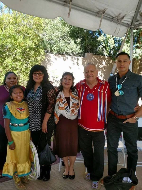 A group of six Indigenous people stand together under a white tent outdoors, including a smiling girl in a yellow and teal dress holding hoops, women, an elder in a red ribbon shirt, and a young man with turquoise jewelry.