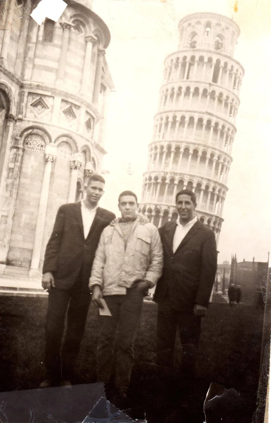 Vintage black-and-white photo of three young men standing on grass before the Leaning Tower of Pisa and the cathedral; two wear dark suit jackets, the one in the middle a light jacket.