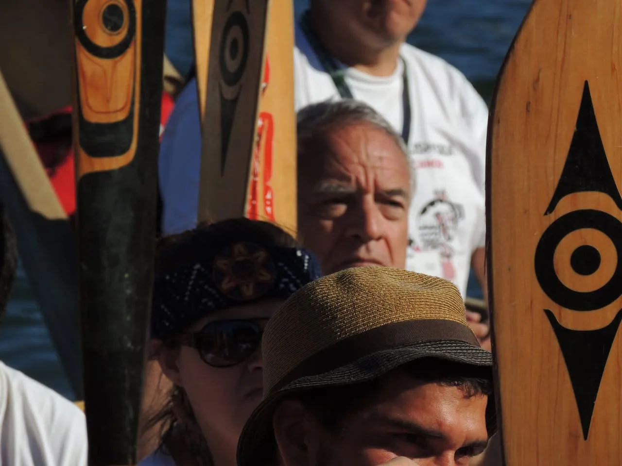 People gathered closely among upright wooden canoe paddles painted with black Northwest Coast formline designs, with water in the background.