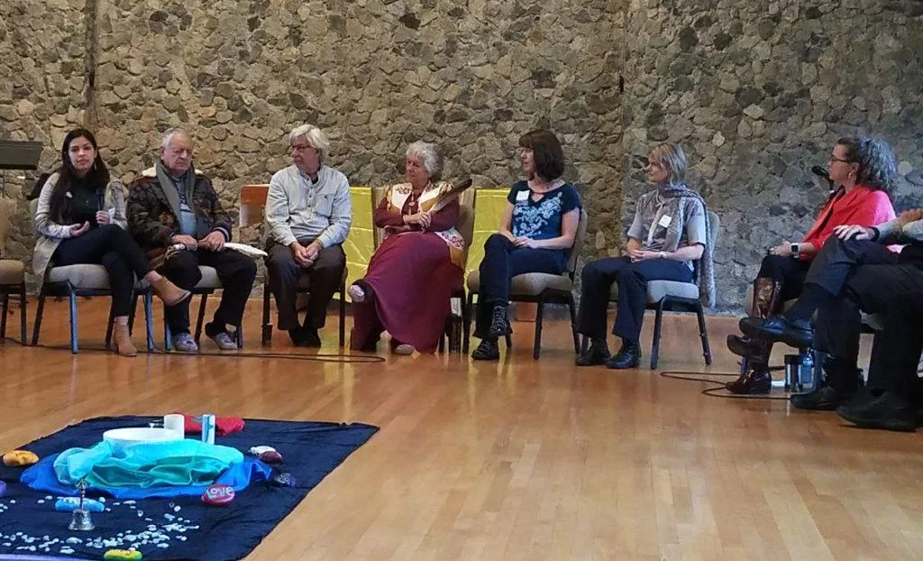 A panel of seven people seated in a semicircle of chairs before a stone wall, one woman speaking into a microphone, with a blue cloth centerpiece holding a bowl, stones and a bell on the wooden floor.