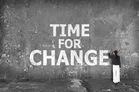 A person in dark clothing stands beside a large gray wall with the words "TIME FOR CHANGE" written in white.