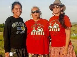 Three women stand together outdoors on a grassy field; an elder with white hair and sunglasses in a red shirt is flanked by a young woman in black and a woman in a red shirt and hat.
