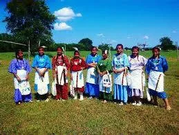 A row of young women in traditional dresses, many blue and some red, with decorated aprons, stand on a green field with trees behind them.