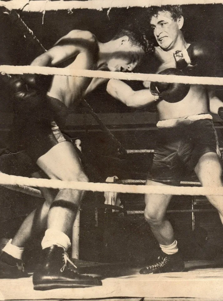 Black-and-white photo of two boxers exchanging punches in a ring, one leaning forward into the ropes.