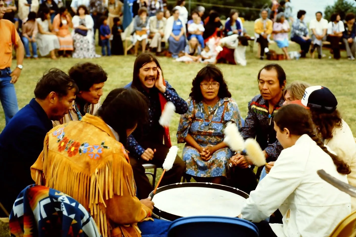 Singers seated around a large drum on a grassy field, raising padded drumsticks as one man sings with a hand to his ear, while a crowd watches seated in a circle behind them.