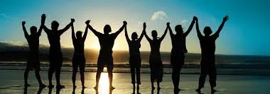 Silhouettes of a row of people holding hands with arms raised, standing on a beach at sunset with the sun glowing behind them.