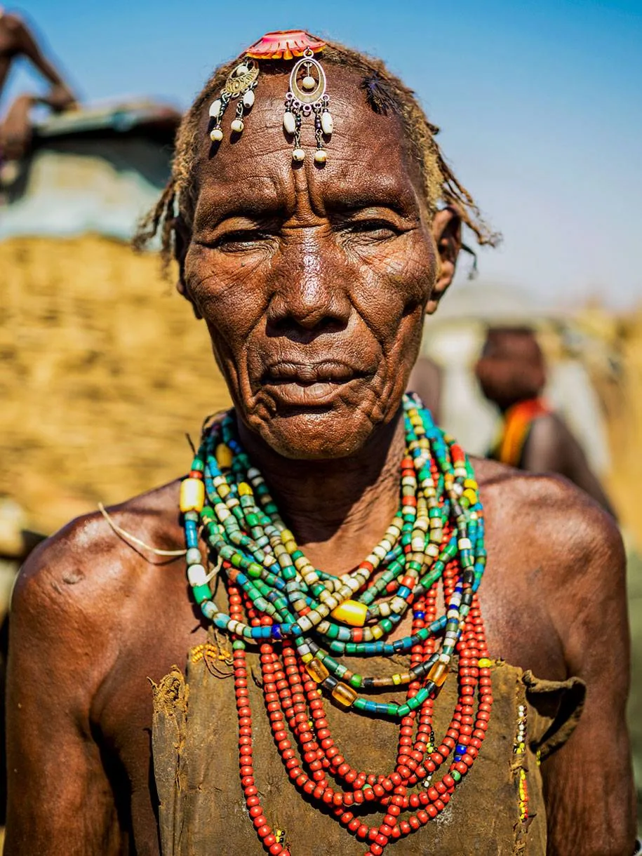 An elder woman with deeply lined skin wears layers of colorful beaded necklaces and a beaded ornament on her forehead, outdoors under a blue sky.